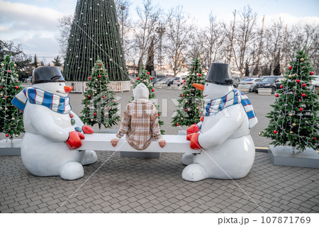 Woman Christmas Square. Close-up of a winter white toy snowman With trees decorated with Christmas tinsel in the background Woman Christmas Square. Close-up of a winter white toy snowman With trees decorated with Christmas tinsel in the background 107871769