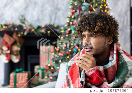Freezing man sitting under a blanket at home on the sofa in the living room, the heating is turned off, the heating of the apartment is not working, hispanic people for Christmas near the tree. 107872364