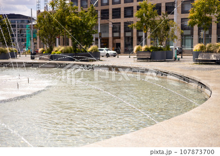 Fountain in the summer city park. Water jets in the fountain. A beautiful park with fountains and trees against of buildings 107873700