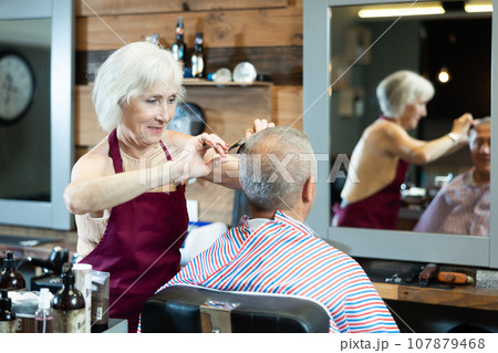 Experienced female hairdresser cutting hair of elderly client with scissors in barbershop Experienced female hairdresser cutting hair of elderly client with scissors in barbershop 107879468