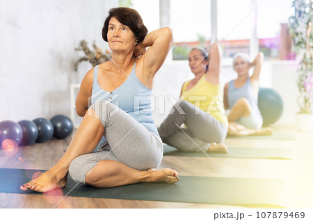 Old woman practicing cow face pose of yoga in training room during fitness classes Old woman practicing cow face pose of yoga in training room during fitness classes 107879469
