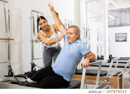 Smiling personal female trainer assisting mature man exercising on Pilates machine in rehabilitation center Smiling personal female trainer assisting mature man exercising on Pilates machine in rehabilitation center 107879523