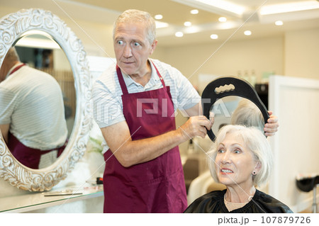 Hairdresser elderly man holds mirror behind senior female client and shows result of haircut. Hairdresser elderly man holds mirror behind senior female client and shows result of haircut. 107879726
