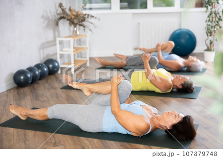At yoga class elderly women is lying on back, bent one leg at knee and raised it to chest 107879748