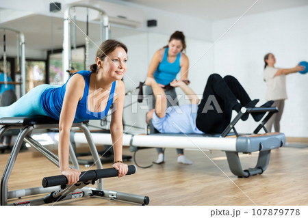 Positive middle-aged woman performing stretching exercises while using pilates Wanda chair machine in rehabilitation center 107879778