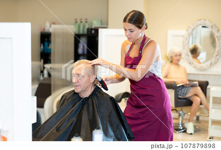 Young woman hairdresser performs haircut for elderly man using hair clipper 107879898