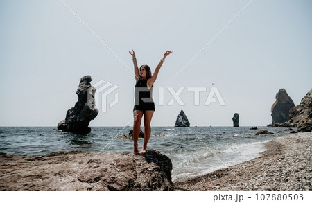 Woman summer travel sea. Happy tourist in hat enjoy taking picture outdoors for memories. Woman traveler posing on the beach at sea surrounded by volcanic mountains, sharing travel adventure journey 107880503