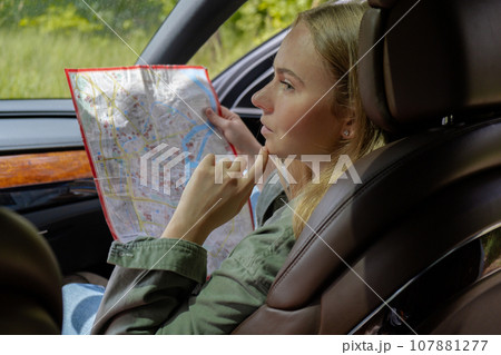 Blonde woman sitting inside the car checking the route on map to get to the destination. Afraid to get lost in forest. Young tourist explore local travel making candid real moments. True emotions 107881277
