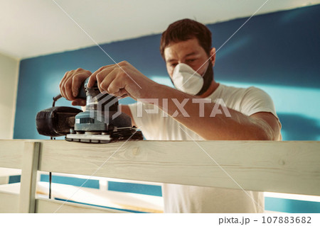 man rubs wooden child bed with power sander or grinder removing old paint 107883682