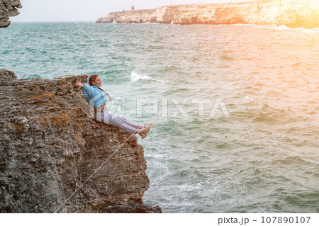 woman sea travel. A woman in a blue jacket sits on a rock above a cliff above the sea, looking at the stormy ocean. Girl traveler rests, thinks, dreams, enjoys nature 107890107
