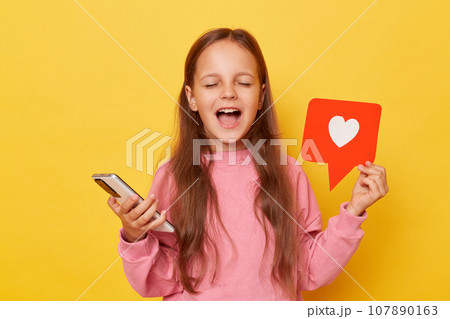 Excited amazed little girl sharing popular content on social media showing blogger heart like icon and using smartphone yelling with closed eyes rejoicing isolated over yellow background 107890163