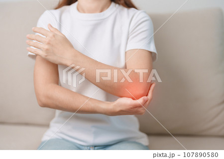 Woman having elbow ache during sitting on couch at home, muscle pain due to lateral epicondylitis or tennis elbow. injury, Health and medical concept Woman having elbow ache during sitting on couch at home, muscle pain due to lateral epicondylitis or tennis elbow. injury, Health and medical concept 107890580
