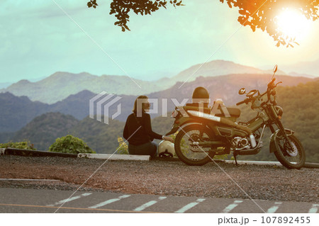 woman sitting beside motorcycle at beautiful viewpoint and looking to sunset scene 107892455