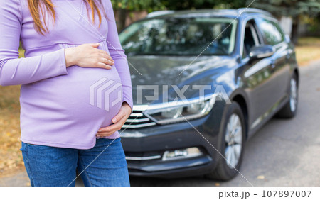 A pregnant girl with a big belly in a purple sweater on the background of a car. The concept of travel and a long trip in a car during pregnancy. Copy space for text 107897007