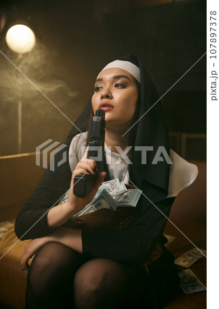 Portrait of attractive nun blowing on handgun barrel after shot 107897378