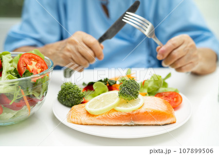 Asian elderly woman patient eating Salmon steak breakfast with vegetable healthy food while sitting and hungry on bed in hospital. 107899506