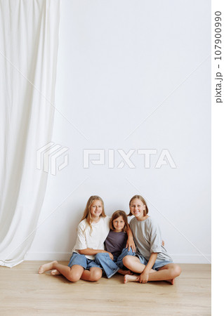Three girls sisters sitting on the floor against a white wall Three girls sisters sitting on the floor against a white wall 107900990