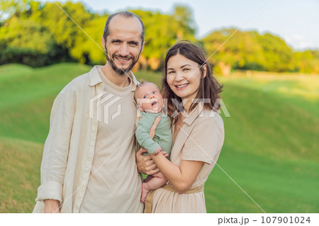 A happy 40-year-old couple cradles their newborn in a sun-drenched park. Love, family and generations in harmony 107901024