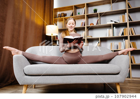 Young beautiful blonde woman in brown leggings and top sitting on a twine at home on the couch. The girl is reading a book sitting on the couch in an unusual position Young beautiful blonde woman in brown leggings and top sitting on a twine at home on the couch. The girl is reading a book sitting on the couch in an unusual position 107902944