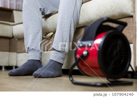 Frozen feet of a man in socks sitting on the sofa are warmed up under the warm air of an electric heater that stands on the floor in a cold apartment, focus on the feet Frozen feet of a man in socks sitting on the sofa are warmed up under the warm air of an electric heater that stands on the floor in a cold apartment, focus on the feet 107904240