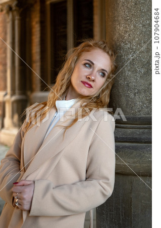 Stylish elegant young woman in beige trench coat with long red hair outside near gray column looking into the distance. Vertical frame. 107904684