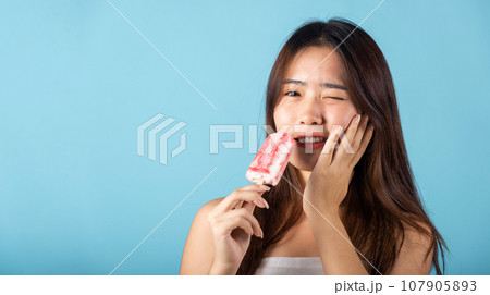 Portrait of Asian young woman with sensitive teeth after eating delicious ice cream wood stick mixed fruit flavor feeling painful uncomfortable, studio shot isolated on blue background, dental problem 107905893