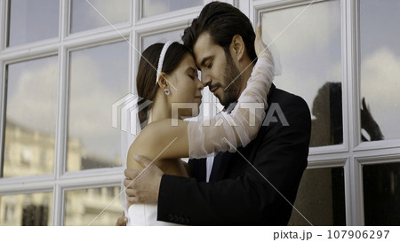 Portrait of loving embracing newlyweds in wedding suit and dress. Action. Tender loving new family in front of windows. 107906297
