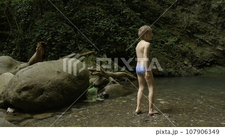 Boy walking in the forest with naked feets in the water. Creative. Boy in summer stream enjoying beautiful landscape. 107906349
