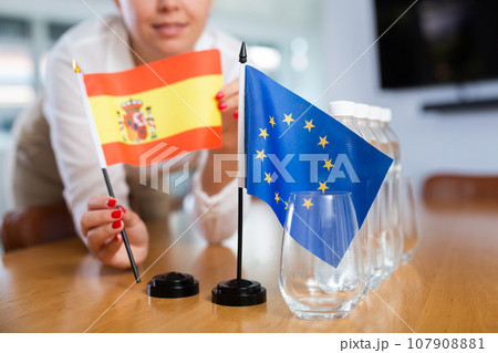 Young woman puts Spain and EU flags on table in office 107908881
