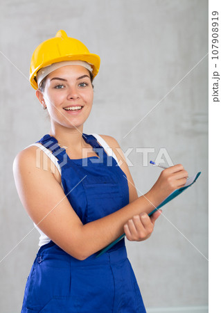 Portrait of emotional mechanic girl in overalls and a protective helmet, who writes in documents 107908919