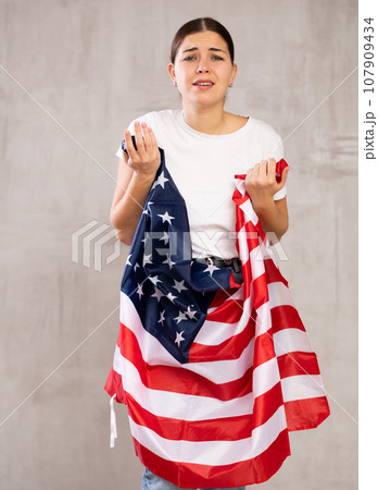 Sad young woman holding USA flag against unicoloured background 107909434