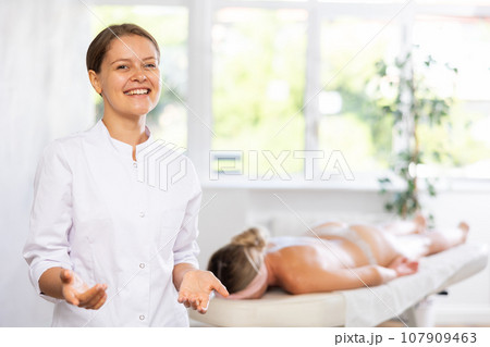 Positive young professional masseuse standing in massage room Positive young professional masseuse standing in massage room 107909463