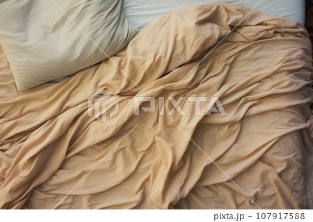 Top view of an unmade beige bed with a crumpled sheet, blanket and pillows 107917588