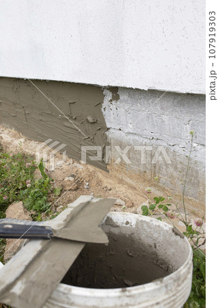 Vertical photo of a bucket with cement and a spatula against the background of the foundation of a house. 107919303