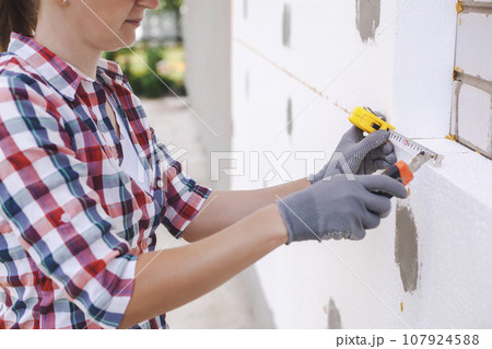 Insulation of the house with polyfoam. The worker is checking with the construction level the accuracy of the installation of polystyrene board on the facade. Insulation of the house with polyfoam. The worker is checking with the construction level the accuracy of the installation of polystyrene board on the facade. 107924588