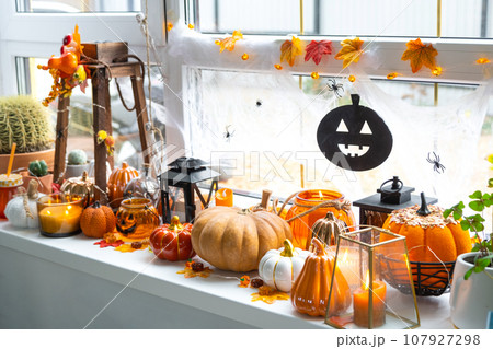 Festive decor of the house on the windowsill for Halloween - pumpkins, Jack o lanterns, skulls, cobwebs, spiders, skeletons, candles and a garland - a cozy and terrible mood 107927298