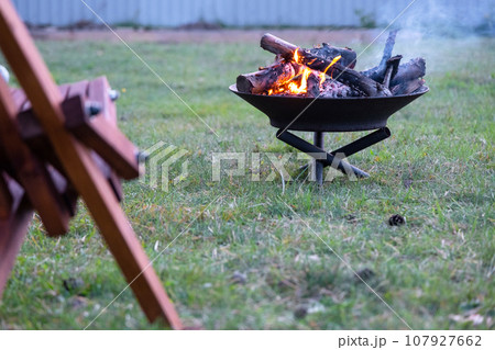 Metal fire bowl with burning wood in the yard and garden rack deck chairs. Safe decorative fire pit, Picnic area in the garden 107927662