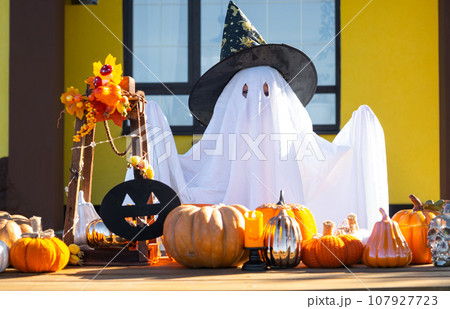 Child in bed sheet with slits like ghost in witch's hat and Halloween decor on the porch of the house outside in the yard of pumpkin, lantern, garlands, ack lantern. party, autumn mood Child in bed sheet with slits like ghost in witch's hat and Halloween decor on the porch of the house outside in the yard of pumpkin, lantern, garlands, ack lantern. party, autumn mood 107927723