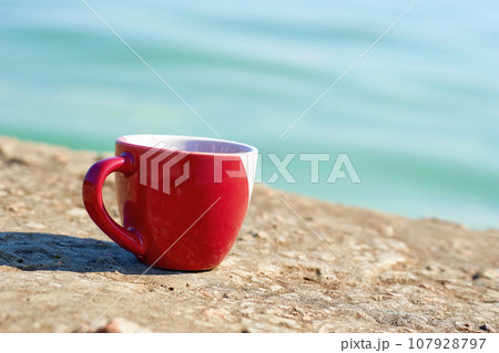 Red ceramic cups, coffee tea beverages on the shore of a stone rocky sea bay  107928797