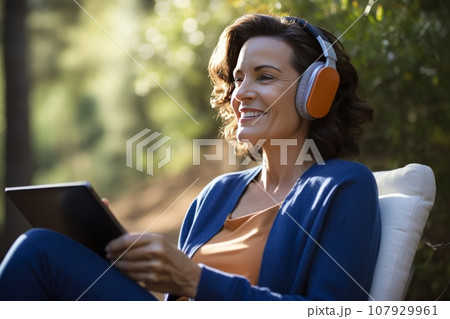 Charming woman wearing headphones and holding tablet sits in a comfortable armchair at country house. Middle-aged caucasian lady listens to music and browses the Internet in the silence of her garden. 107929961