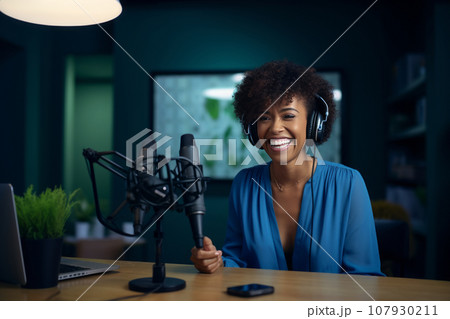 Young beautiful African American woman wearing headphones in a recording studio. Smiling black lady recording news, podcast or blog using microphone, laptop and professional studio equipment. 107930211