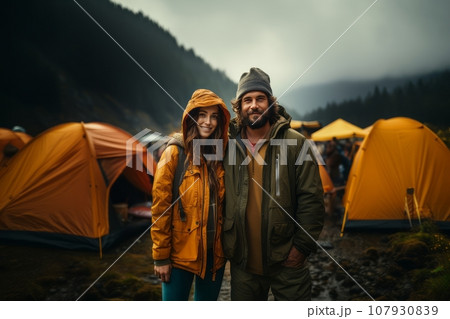 Portrait of happy young couple in the mountains against the backdrop of a tent camp. Caucasian family in outwear is engaged in a mountain hike. Active lifestyle, tourism and vacation concept. 107930839
