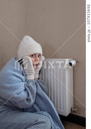 a warmly dressed woman sits near a radiator and is shocked by the lack of heating 107933698