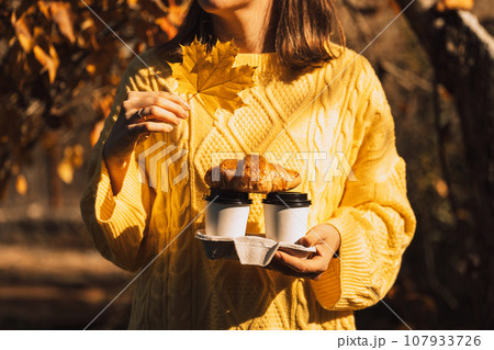 Lady in yellow knitted sweater holding coffee to go and croissant in front of yellow leaves 107933726