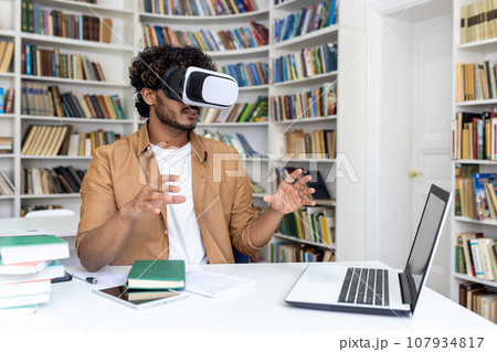 Young hispanic male student sitting at a desk in a university library, studying online on a laptop wearing a virtual mask. Gesturing and waving his hands. 107934817
