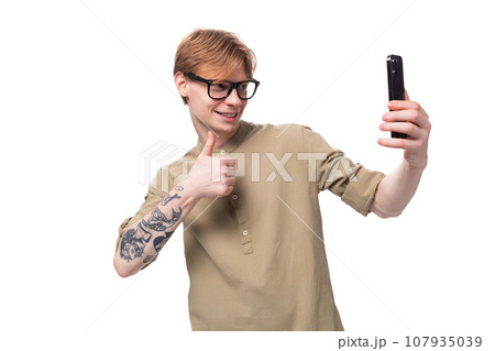a young red-haired guy in glasses and a brown shirt takes a selfie on the phone 107935039