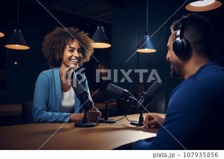 Happy smiling multiethnic blogger couple hosting a podcast in the studio. Cheerful woman and man wearing headphones sit at a table with microphones and chatting cheerfully. Modern media technologies. 107935856