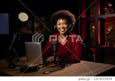 Young beautiful African American woman wearing headphones in a recording studio. Smiling black lady recording news, podcast or blog using microphone, laptop and professional studio equipment. 107935934