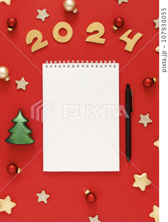 White sheet of notebook on red table with golden stars, Christmas balls, yellow numbers of 2024 . Concept of planning, wish list for the new year 107938055