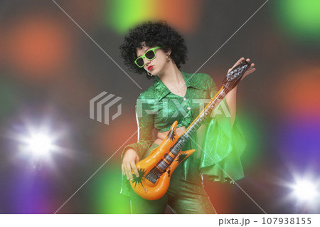 Retro disco woman with comic guitar posing against colorful stage lights. 107938155